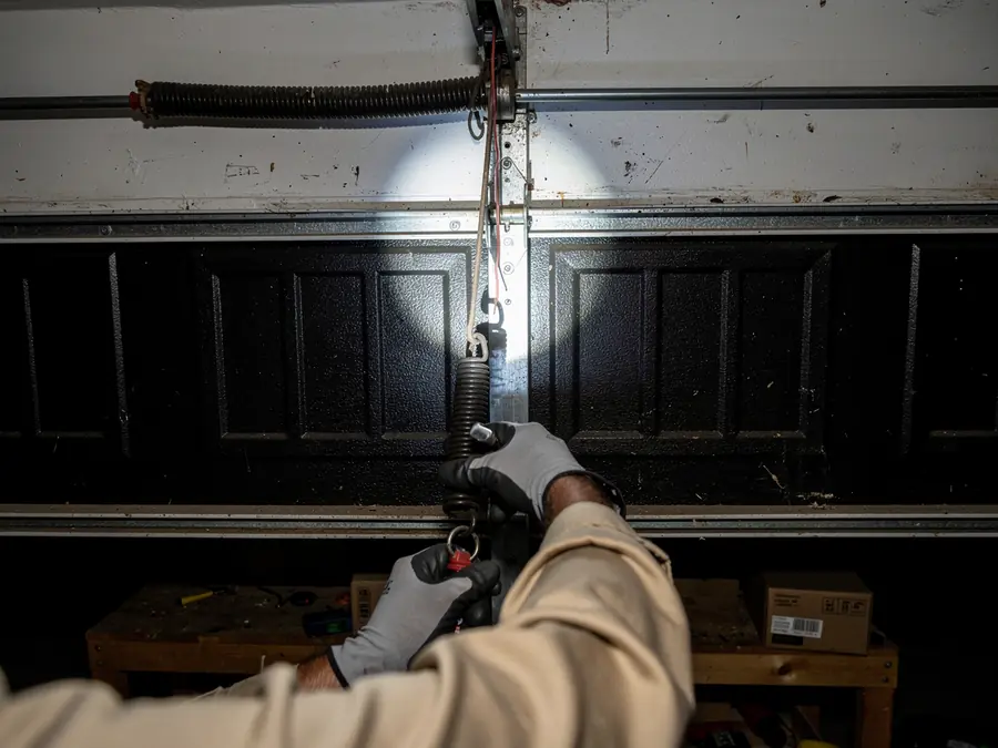 A technician in gloves works on a garage door spring and cable.